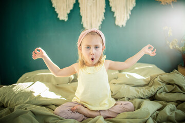 Little caucasian girl playing yoga and has fun on the bed in a bright sunny room