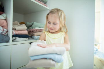 Little caucasian blonde girl neatly folds clothes on her shelf
