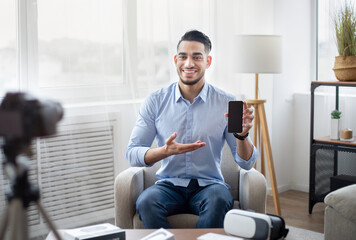 Arab male influencer filming video review of contemporary smartphone on dslr camera at home, mockup on screen