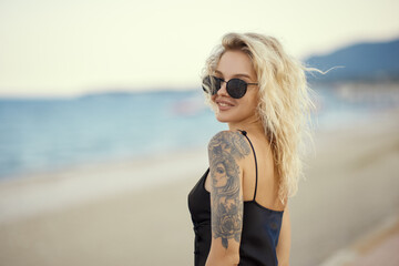 Portrait of gorgeous blonde woman in summer sexy black dress and sunglasses relaxing outdoors against beach on the background. A fashionable happy young adult lady standing at the sea coast looking to - Powered by Adobe
