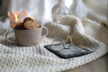 Bowl of chocolate chip cookies, digital tablet, reading glasses, soft blanket and lit candle. Hygge at home. Selective focus.