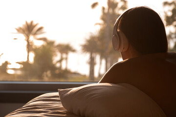 Silhouette of woman with headphones hugs pillow lying on the bed in the bedroom opposite panoramic window with view on palm tree sea beach at sunset. Female relaxing listening music.