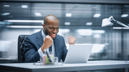 Office: Happy Successful Black Businessman Sitting at Desk Using Laptop Computer, Celebrate Success. Entrepreneur in Suit working with Stock Market App Smiles, Happy Victory. Motion Blur Background.