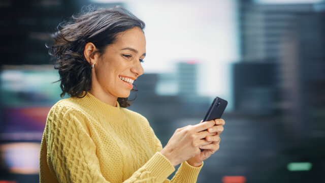 Street Shot: Portrait of Beautiful Latin Woman Using Smartphone. Smiling Hispanic Female Entrepreneur Using Mobile Phone for Online Shopping, e-Commerce Happily. Blurred Motion Background.
