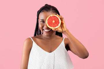 Fruit vitamins and detox. Cheery black woman with afro bunches covering eye with grapefruit half on pink background