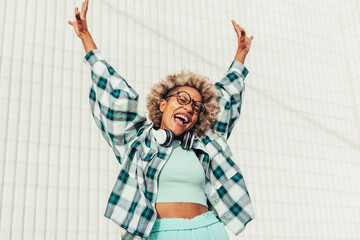 Cheerful young carefree afro woman with arms raised and smiling outside listening music. Happy latin american female celebrating freedom outdoors.