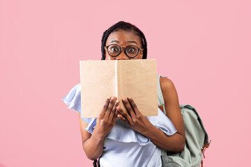 Shocked African American female student with backpack hiding behind open book on pink studio background