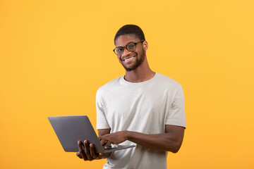 Excited black freelancer man using laptop, standing in studio over yellow background and smiling to...