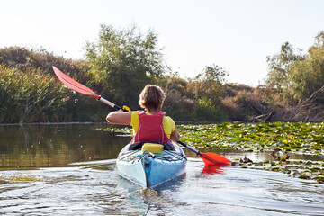 Rear view of woman paddle in blue kayak in the small river among the water lilies and driftwoods