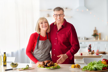 Cheerful senior couple posing, cooking turkey for Christmas or Thanksgiving dinner, preparing holiday meal in kitchen