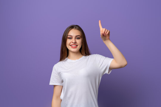 Happy young woman with long hair pointing finger up, raising eyebrows up surprised, showing advertisement, standing over purple background