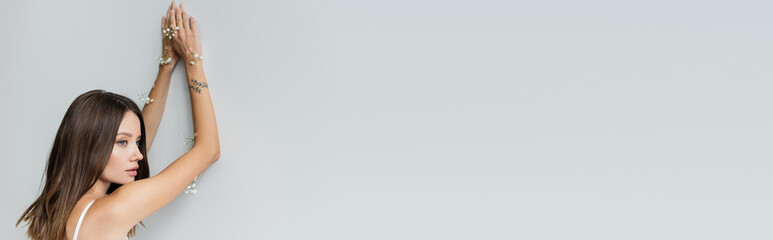 Fototapeta premium young tattooed woman with white gypsophila flowers on body looking away on grey background, banner.