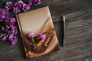 small purple carnations are laid out in the corners on a wooden gray background, where lies a notebook and a pen