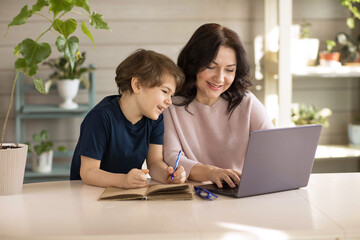 Mother and son are engaged in online school, sit in a laptop and make notes in a notebook