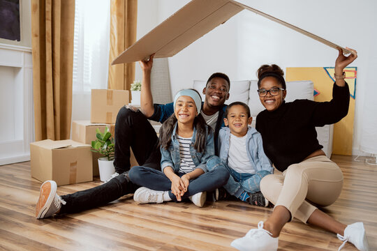 Family complete, parents with two children sitting on the floor relaxing after moving to a new apartment, holding over their heads a piece of cardboard left from boxes as a roof, smiling at the camera