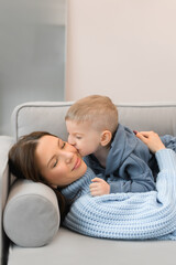 Fototapeta premium A young mother and a little son spend time at home. They lie in the living room on the sofa and hug, kiss.