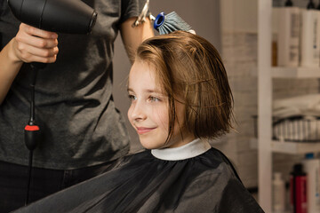 Hairdresser blow-dry model short wet hair after cutting. Satisfied Child looks in mirror and admires new image. Short-haired little girl makes smiles in barbershop