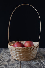 Wicker basket with ripe seasonal red apples, wet after washing with the remaining drops of water, ready to eat, placed on a rustic wooden table against a black wall, space for text.