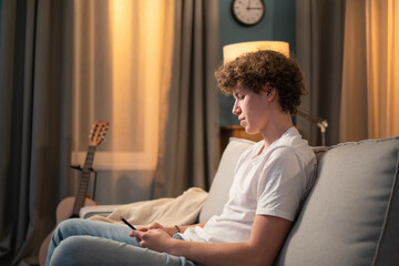 A bored young boy, teenager, schoolboy, student is relaxing on the couch in the evening with his phone, smartphone in hand. The young boy sends messages to friends, browses social media