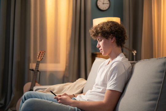 A bored young boy, teenager, schoolboy, student is relaxing on the couch in the evening with his phone, smartphone in hand. The young boy sends messages to friends, browses social media