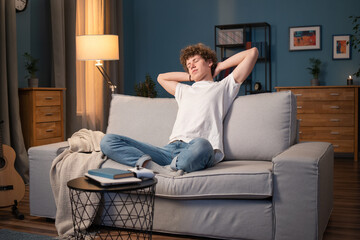 A young student sits on the living room couch in the evening with legs crossed. The teenager stretches out his arms, puts his hands behind head. Collage Boy sits with eyes closed