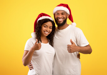 Happy New Year. Cheerful black spouses in Santa hats showing thumbs up and smiling to camera on yellow background