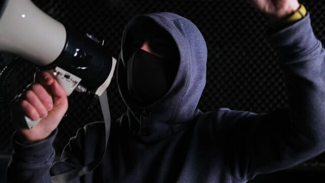 A male activist in a hood and a black mask shouts slogans into a megaphone and shakes his hand. Protest. The concept of an uprising, a rally and the overthrow of power, the defense of rights