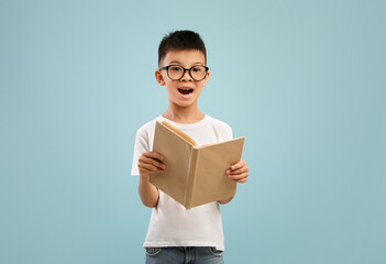 Excited little asian boy wearing eyeglasses holding and reading open book