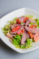 Healthy green salad with meat, tomato and lettuce served in a square white plate over grey or gray background.
