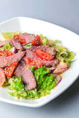 Healthy green salad with meat, tomato and lettuce served in a square white plate over grey or gray background.