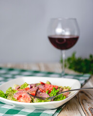 Healthy green salad with meat, tomato and lettuce served in a square white plate with a glass of red wine.