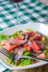 Healthy green salad with meat, tomato and lettuce served in a square white plate with a glass of red wine.