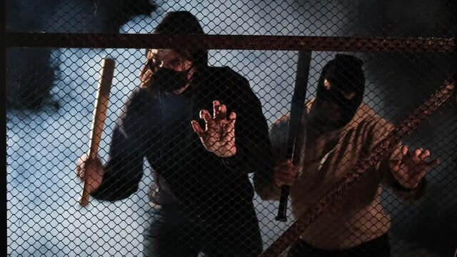 Two aggressive masked men with baseball bats are trying to break down the fences. Smoke in the background. The concept of pogroms and protests, uprisings and overthrow of power