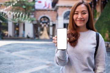 Mockup image of a young asian woman holding and showing a mobile phone with blank white screen while traveling in the city