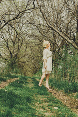 Naklejka na ściany i meble Beautiful young woman in white chiffon dress with short blonde haircut walking along barefoot the path and looking at the trees