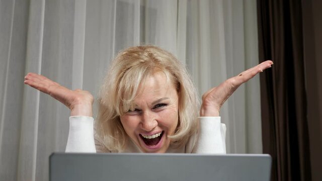 Blonde long-haired woman of middle age wearing warm white turtleneck smiles happily after reading good news using internet on laptop closeup.