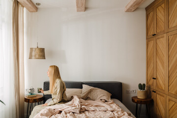 Young blonde woman drinking water while sitting on bed after sleep
