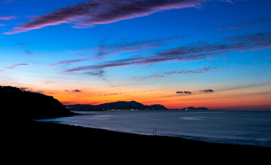 Vista de la playa de Azkorri al anochece sobre el mar con cielo azul con horizonte amarillo anaranjado y nubes rosáceas con el sol iluminando entre las nubes en el centro de la foto