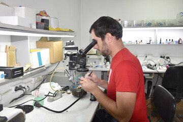 A young man in the workshop examines the microcircuit of an electrical device through a microscope, performing repairs with a screwdriver and scalpel