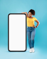 Joyful Afro woman in casual wear leaning on huge cellphone with empty white screen on blue background, mockup