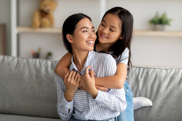 Happy asian little girl hugging her beautiful mother and smiling to each other, sitting on sofa in living room