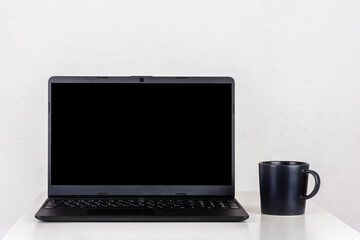 a black mockup on a laptop screen and a black mug of tea on a table against a white wall with a copy space