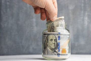 Hand puts american dollar banknotes in a glass jar, saving money concept, dark background with copy space