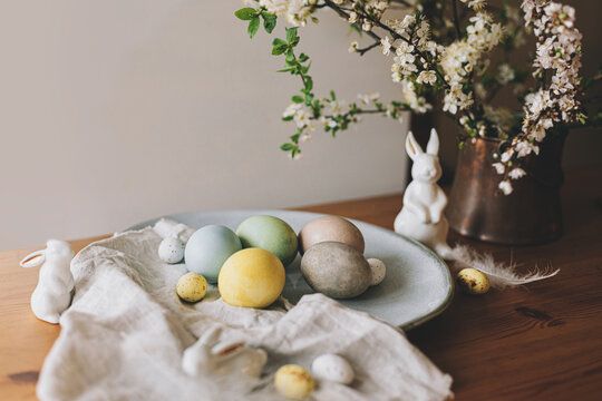 Easter eggs on plate with bunny figurines, linen napkin,  cherry blossoms on rustic table. Happy Easter! Natural dyed colorful eggs and spring flowers. Countryside still life