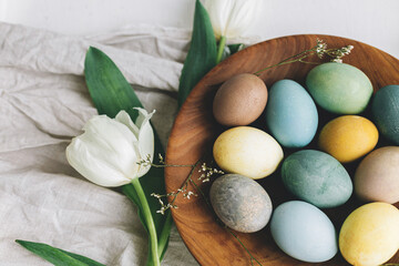 Stylish Easter eggs in wooden plate, tulips and linen napkin on rustic white table. Happy Easter! Natural dyed colorful eggs and spring flowers rustic composition.
