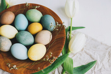 Happy Easter! Stylish Easter eggs in wooden plate, tulips and linen napkin on rustic white table.  Natural dyed colorful eggs and spring flowers rustic composition.