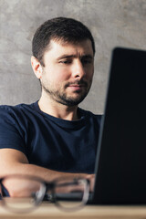 Handsome serious bearded man sitting at the office desk with laptop against concrete wall in modern office
