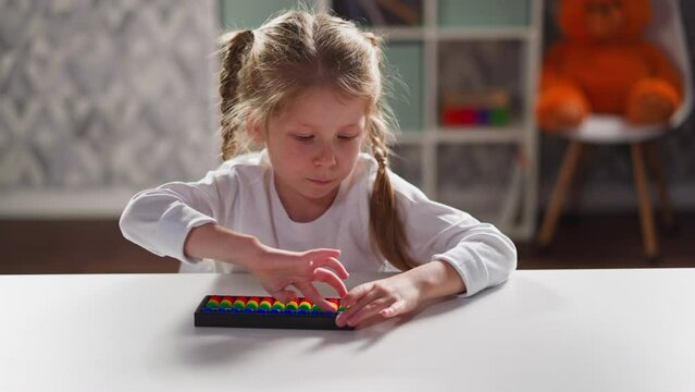 Pretty little girl student learns to solve tasks using toy abacus sitting at white desk during arithmetic lesson at home closeup slow motion