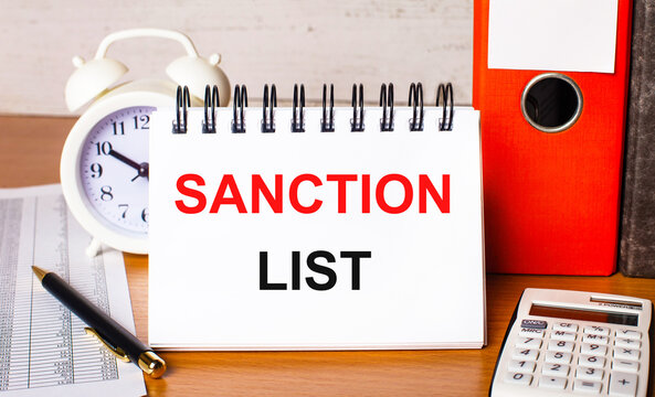 On the table are reports, a white alarm clock, a calculator, folders for papers, a pen and a white notebook with the SANCTION LIST. Business concept