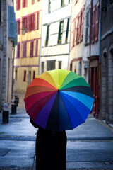 Una persona con ropa negra y un paraguas con los colores del arcoiris de espaldas paseando por una calle de un pueblo frances